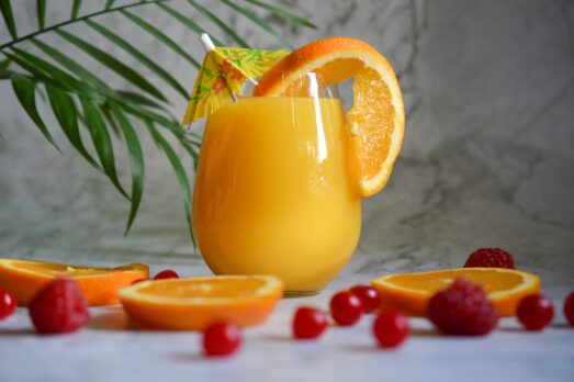 Świeży sok pomarańczowy wyciskany bezpośrednio do szklanki za pomocą wyciskarki do cytrusów zamontowanej na Thermomixie, otoczony przez świeże pomarańcze i maliny