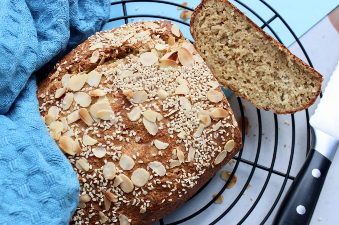 Chleb twarogowy z Thermomix posypany płatkami migdałowymi i sezamem, z jednym przekrojonym kawałkiem ukazującym puszysty miąższ. Chleb spoczywa na metalowej kratce, a w tle widać błękitny materiał o fakturze