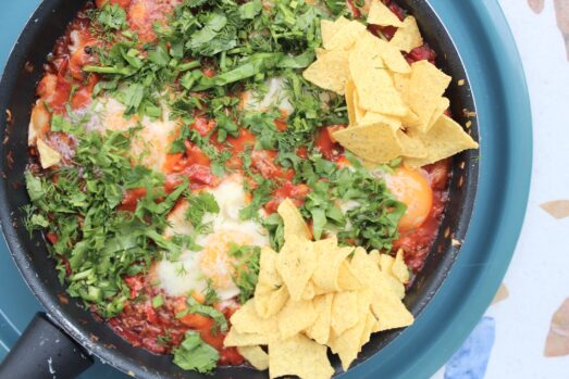 Kolorowe Huevos Rancheros: jajka sadzone na pikantnym sosie pomidorowym z czerwoną papryką, posypane świeżą zieleniną i nachosami