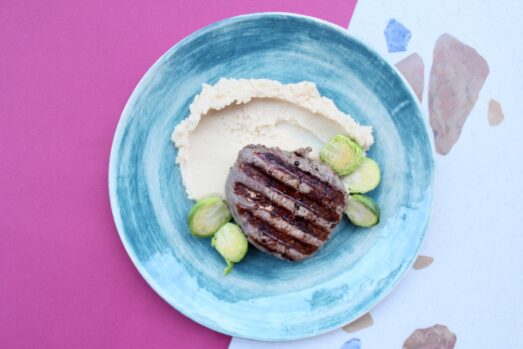 Na zdjęciu widać talerz z posiłkiem umieszczonym na różowym tle z kawałkami niebieskiego i różowego papieru. Talerz ma intensywny, błękitny kolor z efektem przejścia kolorów na wzór akwareli. Posiłek składa się z dobrze upieczonego steka z charakterystycznymi prążkami od grillowania, porcji puree z selera oraz kilku ugotowanych brukselki. Całość prezentuje się apetycznie i jest estetycznie podana.