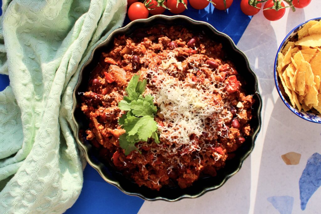 chili con carne przepis z Thermomixa prezentowany za pomocą zdjęcia kolorowej, pikantnej potrawy w ceramicznym naczyniu, posypanej serem i kolendrą, z nachosami i pomidorkami w tle