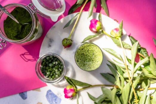 Na obrazku widoczna jest szklanka soku z młodego jęczmienia na białym talerzu, otoczona słoikiem z sproszkowanym sokiem z młodego jęczmienia oraz zielonymi tabletkami. Sok z młodego jęczmienia ma wiele właściwości zdrowotnych i pozytywnych opinii. Dowiedz się, jak pić sok z młodego jęczmienia, na co pomaga i jak zrobić sok z młodego jęczmienia. Możesz także znaleźć informacje, gdzie kupić sproszkowany sok z młodego jęczmienia.